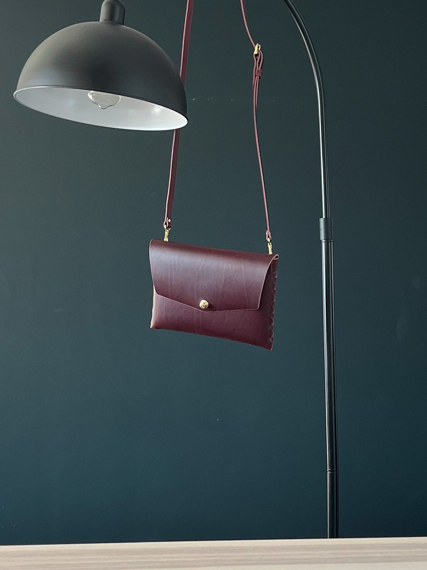 modjūl’s leather mini bag deluxe bag in olive, hanging on a lamp. A larger take on the classic mini bag, a rectangular-shaped bag with long detachable straps, handcrafted in Canada using quality Italian leather and brass hardware.
