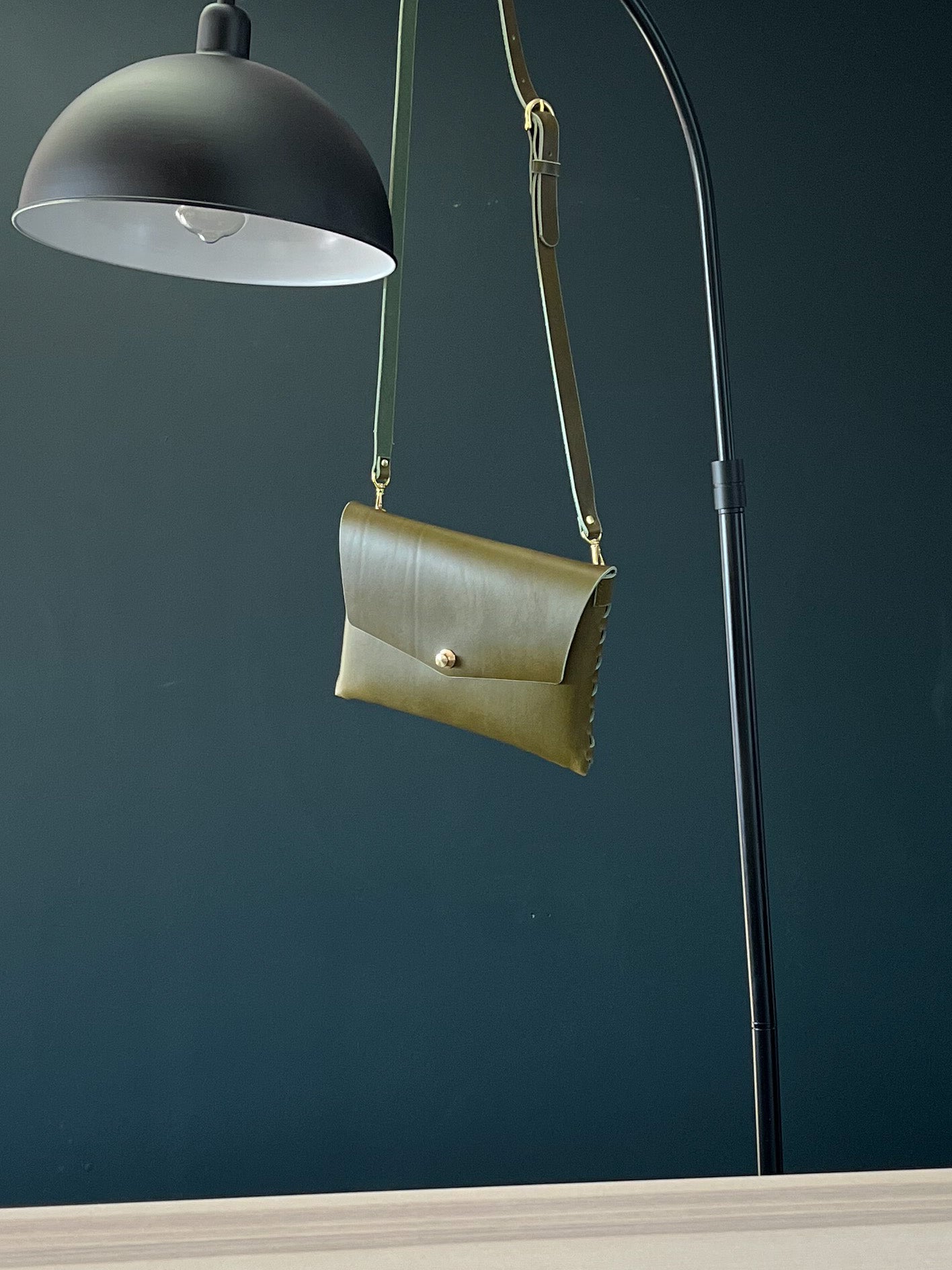 modjūl’s leather mini bag deluxe bag in olive hanging on a lamp. A larger take on the classic mini bag, a rectangular-shaped bag with long detachable straps, handcrafted in Canada using quality Italian leather and brass hardware.
