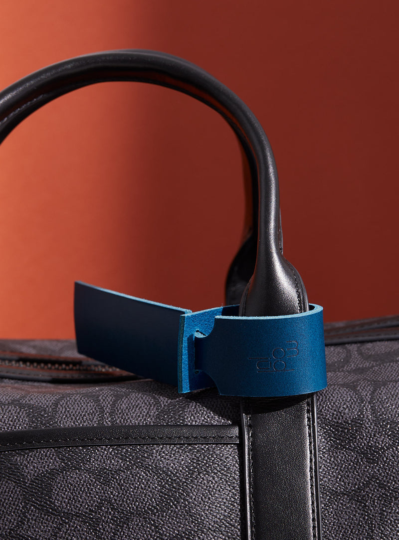 The modjūl leather traveller’s tag in blue, displayed on a duffle bag. A luggage tag constructed using quality vegetable-dyed Italian leather with the option for personalization.