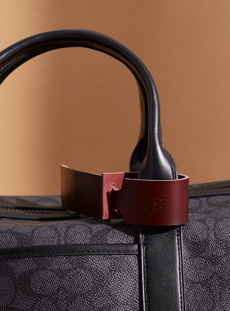 The modjūl leather traveller’s tag in burgundy, displayed on a duffle bag. A luggage tag constructed using quality vegetable-dyed Italian leather with the option for personalization.