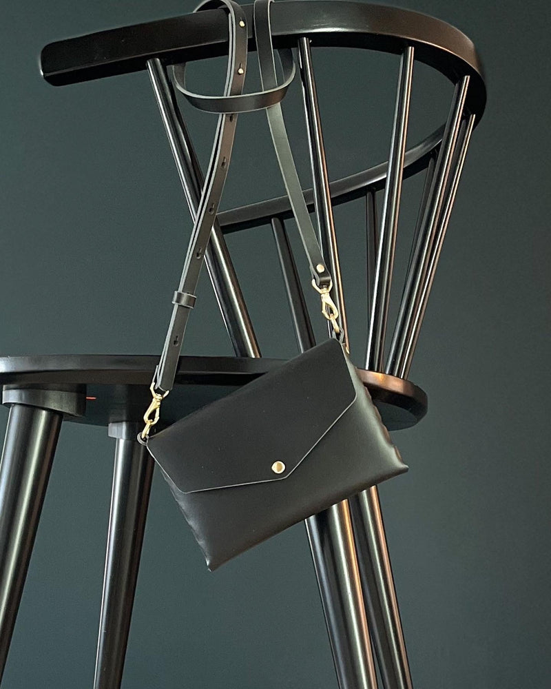 modjūl's mini leather bag in black hanging on a chair.. A rectangular-shaped bag with long straps, handmade in Canada using quality Italian leather and brass hardware.