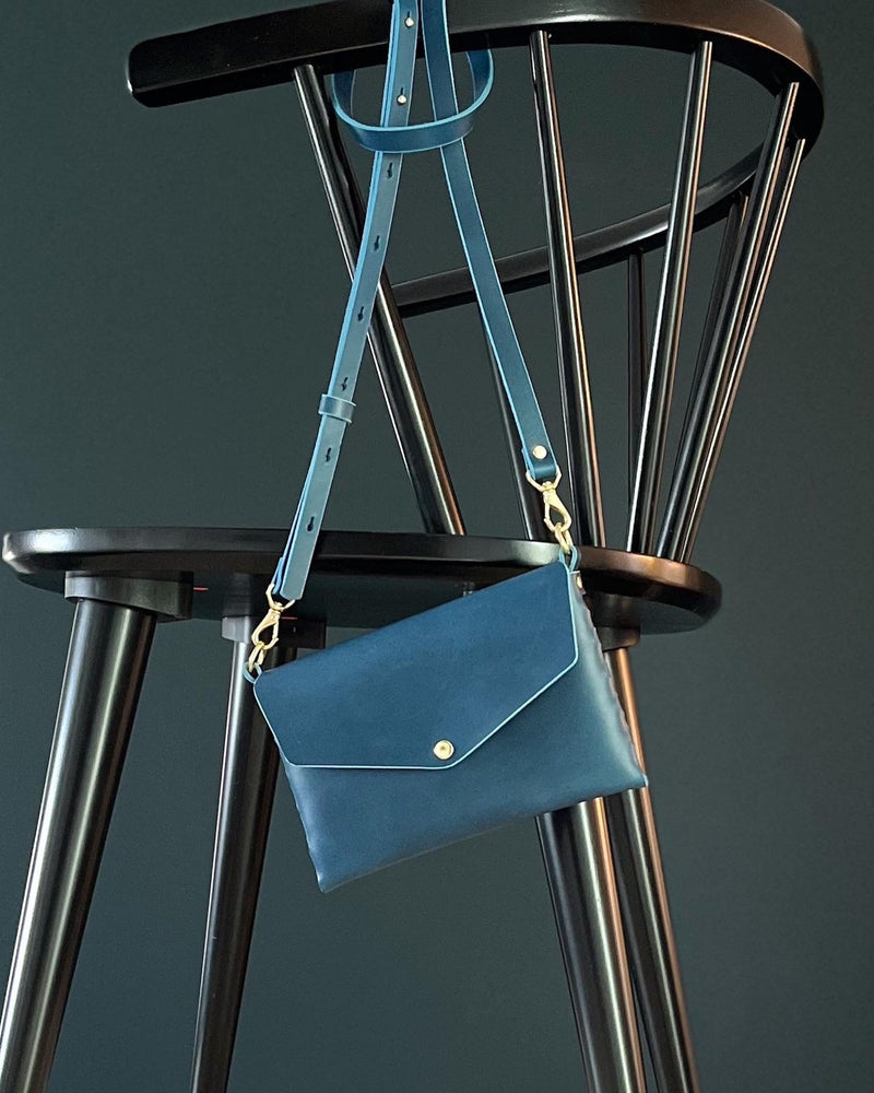 modjūl's mini leather bag in blue hanging on the back of a chair. A rectangular-shaped bag with long straps, handmade in Canada using quality Italian leather and brass hardware.