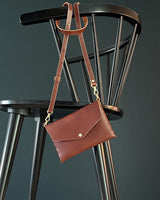 modjūl's mini leather bag in brown hanging on the back of a chair. A rectangular-shaped bag with long straps, handmade in Canada using quality Italian leather and brass hardware.