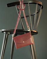 modjūl's mini leather bag in burgundy hanging on the back of a chair. A rectangular-shaped bag with long straps, handmade in Canada using quality Italian leather and brass hardware.