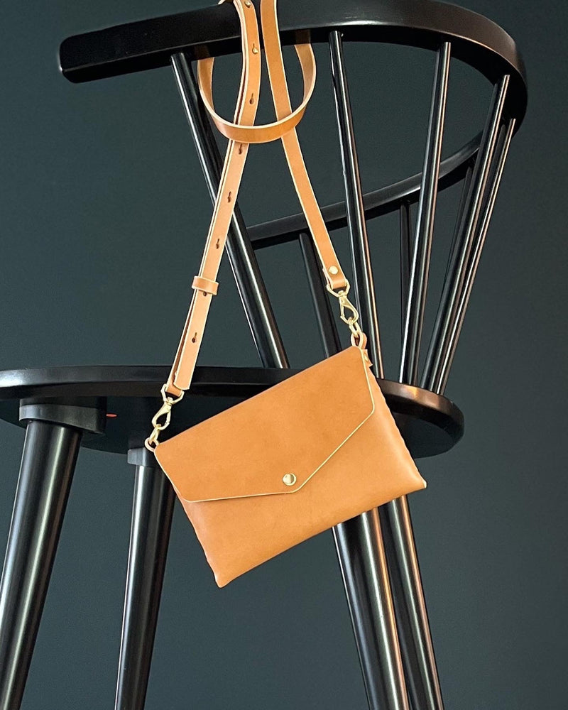 modjūl's mini leather bag in camel hanging on the back of a chair. A rectangular-shaped bag with long straps, handmade in Canada using quality Italian leather and brass hardware.