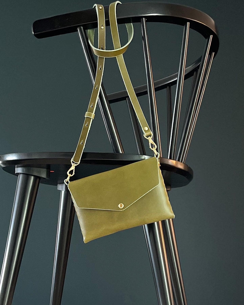 modjūl's mini leather bag in olive hanging on the back of a chair. A rectangular-shaped bag with long straps, handmade in Canada using quality Italian leather and brass hardware.